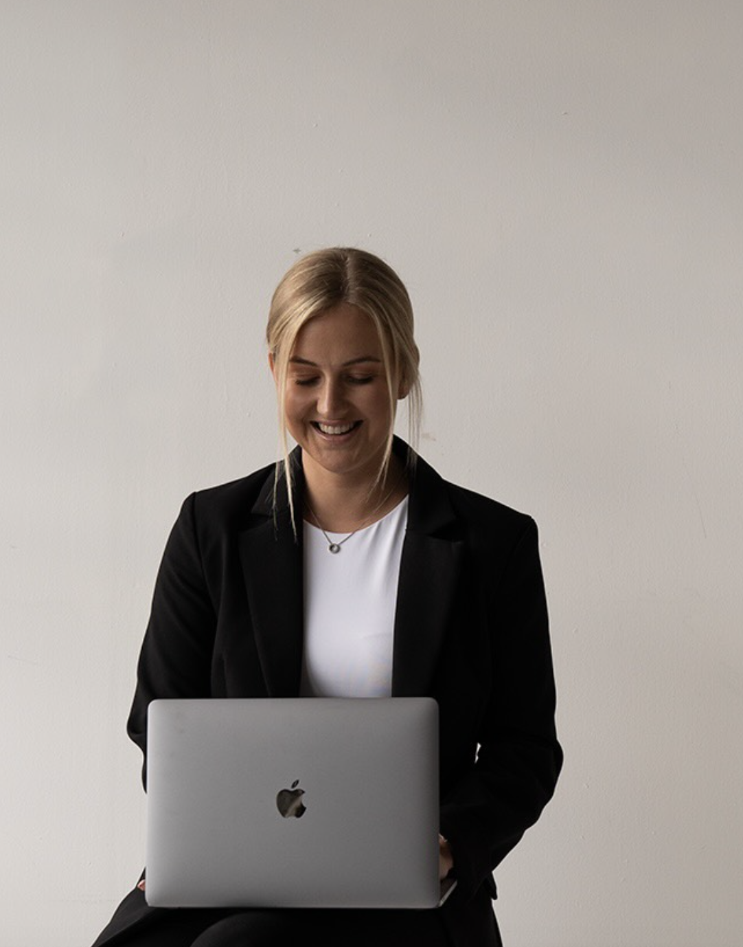 Woman in a black blazer working on a MacBook against a plain wall